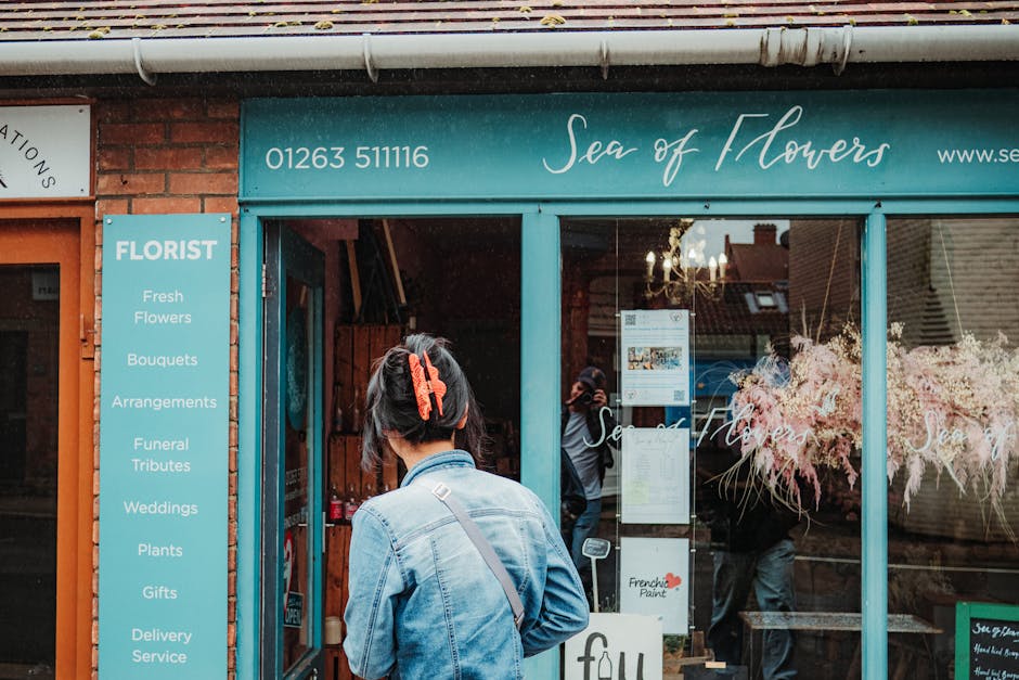 A woman with short dark hair secured with a red hair clip, wearing a light denim jacket and carrying a shoulder bag, stands outside the floral shop 'Sea of Flowers' on Field End Road Eastcote. The sho