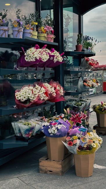 A display of fresh floral arrangements at an outdoor flower stand, featuring vibrant bouquets of white and pink chrysanthemums wrapped in pink and purple tissue paper, along with smaller mixed flower 