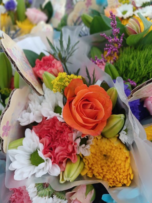 A vibrant bouquet of fresh flowers featuring a prominent orange rose at the center, surrounded by white daisies with green centers, pink carnations, yellow chrysanthemums, and purple statice. The flow