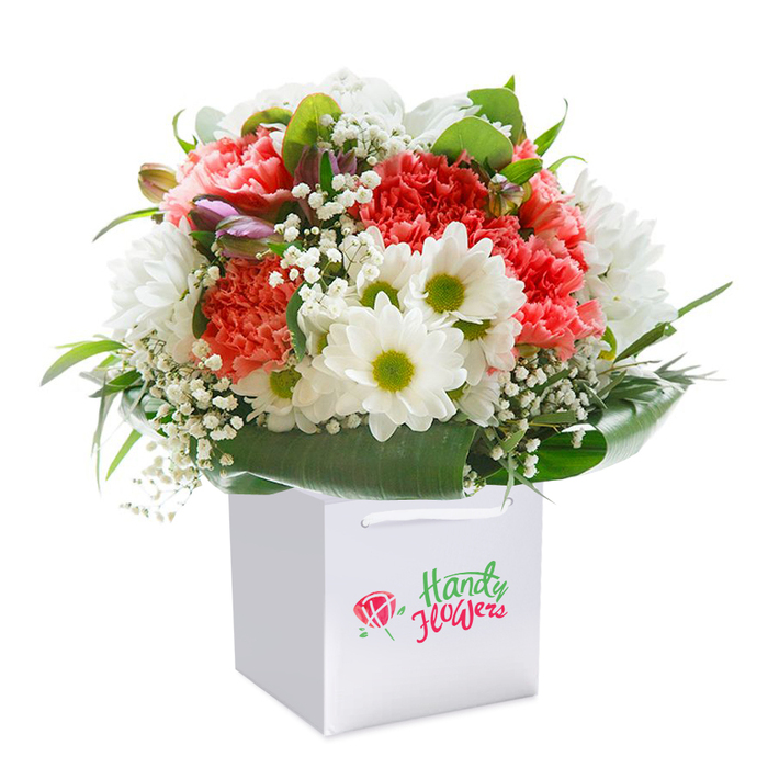Elegant and brimming with fresh energy, this full-bodied bouquet is artfully composed within a stylish square white gift bag-its sleek ribbon handles suggesting easy delivery to a home near Eastcote Village. The visual centrepiece is a generous collection of coral, blush, and peach carnations, each displaying intricate frilled petals whose gradations shimmer beneath gentle, natural light. White, daisy-like chrysanthemums punctuate the bouquet, their clean petals and yellow-lime centers offering a joyful contrast and a sense of openness. Interwoven amongst these, airy clouds of white baby's breath provide textural softness, while on the far left, a few unopened buds tinged with purple add a sense of mystery and anticipation. Lush deep green foliage, marked by broad, glossy leaves, encircles the base of the bouquet-evoking the tranquility of nearby parks and the careful touch of an Eastcote florist. The arrangement as a whole is crafted to form a lush, rounded silhouette, conveying not only the abundance of fresh flowers but also thoughtful, meticulous care. Resting within the crisp white bag, the bouquet is elevated as a gift meant for celebration-maybe marking an important occasion along Field End Road or simply to bring a smile to someone special. The clean white background spotlights every colourful element, making the bouquet the undeniable focal point and a symbol of floristry excellence in the Eastcote area.