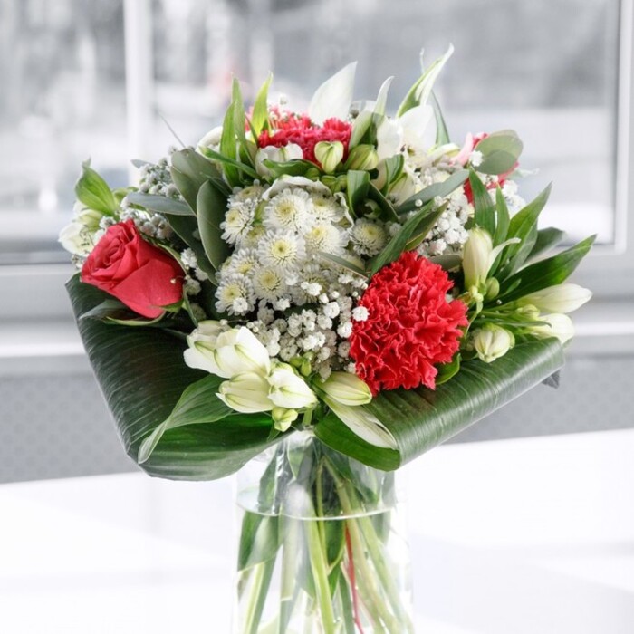 A romantic red and white bouquet is beautifully arranged in a cylindrical glass vase, its clear sides showcasing the carefully trimmed, criss-crossing green stems resting in fresh water. At the forefront, a lush crimson carnation with densely ruffled petals provides a rich textural focal point, backed by velvety deep red roses that are just beginning to open, their layered petals curling outward to suggest unfolding emotion. Interspersed among the passionate reds are clean white and cream flowers: white alstroemeria with their fine speckles and softly flared petals, white chrysanthemums or similar multi-petalled blooms, and several closed white lily buds hinting at future blossoms and lasting beauty. Wisps of gypsophila thread through the bouquet, their tiny white flowers creating a delicate haze that softens the stronger shapes and colours. The entire arrangement is encircled by lush foliage: broad, glossy dark green leaves are artfully wrapped and curled at the base to form a neat, architectural collar, while slimmer, eucalyptus-like greenery and pointed leaves reach upward, introducing varied textures and tones of green. The bouquet rests on a spotless white surface, set against a bright, gently blurred background that feels like daylight filtering through a window in an Eastcote home overlooking a quiet garden. The overall impression is one of balanced harmony and thoughtful craftsmanship, the kind of piece that could easily become the centrepiece of a dining table for a family celebration. Whether delivered as an anniversary surprise, a Mother's Day gesture, or a refined thank-you for a neighbour nearby, the bouquet carries a mood of quiet celebration and heartfelt sincerity, shaped by hands that understand both flowers and the rhythm of local Eastcote life.