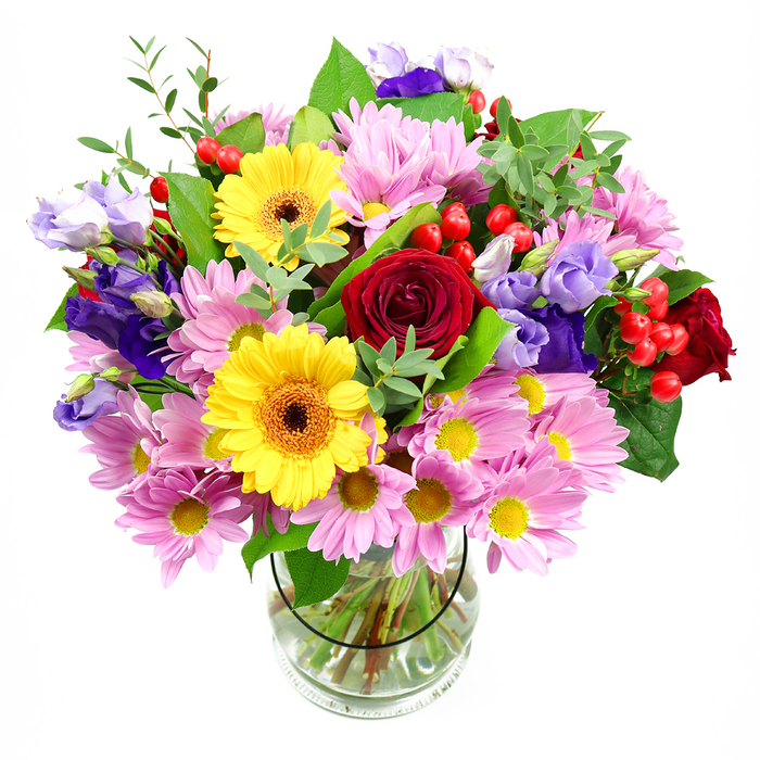 A generous, cheerful bouquet in a clear glass vase is captured against a crisp white background, its colours and shapes arranged with the assurance of a skilled Eastcote florist. At the centre, plush deep red roses bloom in layered spirals, their velvety texture and rich tone lending a sense of heartfelt sentiment. Close by, two radiant yellow gerbera daisies with dark brown-orange centres offer a bright, almost sunburst effect, infusing the design with friendliness and optimism. Clusters of soft pink daisies or chrysanthemums spill outward in gentle domes, their fine, numerous petals and tiny yellow eyes creating a soft-focus frame that cushions the bolder blooms. Threaded between them, stems of purple and lavender lisianthus rise and curve, their bell-shaped blossoms and buds contributing cool, sophisticated notes that echo the calm of nearby green spaces like Pinner Memorial Park. Small sprays of vivid red hypericum berries punctuate the arrangement, their round, bead-like forms adding playful texture and a slightly contemporary twist. Surrounding and supporting the flowers is an array of greenery: some leaves are broad, dark, and glossy, while others are slender and bright, together giving the bouquet natural structure, contrast, and a freshly picked feel. The glass vase reveals neatly gathered stems resting in clear water, a quiet detail that hints at the bouquet's freshness and careful conditioning. This arrangement feels well suited to mark special moments in Eastcote-a birthday celebration, a thank-you gesture delivered to a local office, or a colourful welcome in a home off Eastcote Road-radiating joy, thoughtfulness, and the understated elegance of a handcrafted floral gift.